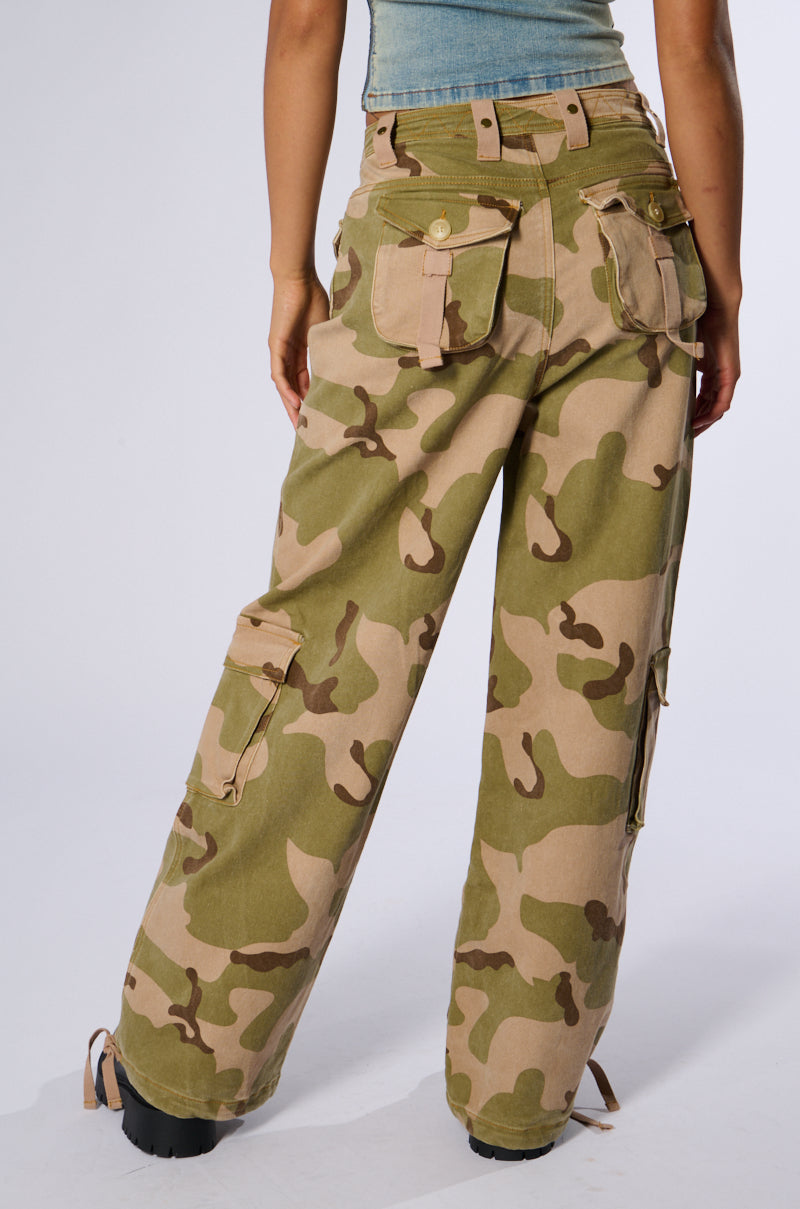 A person wearing the ON TOP OF IT CAMO JOGGER in beige and green camouflage, stands facing away. The joggers feature multiple cargo flap pockets with button closures and a loose, relaxed fit. The background is plain and light-colored.