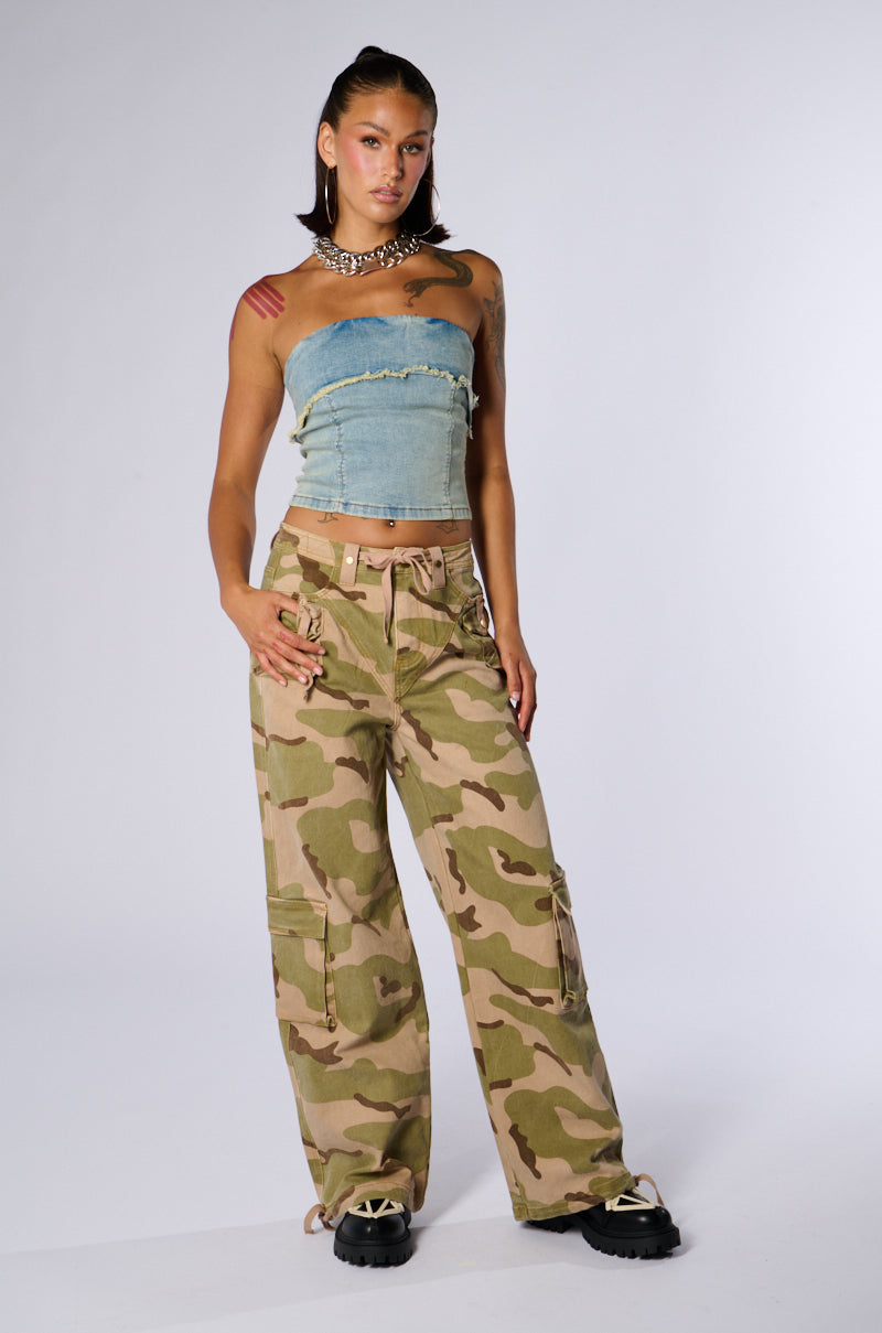 A woman stands against a plain background wearing a strapless, frayed denim crop top paired with the ON TOP OF IT CAMO JOGGER secured with a beige belt. She has her hair pulled back, flaunts a shoulder tattoo, and completes her look with black platform shoes and silver accessories.