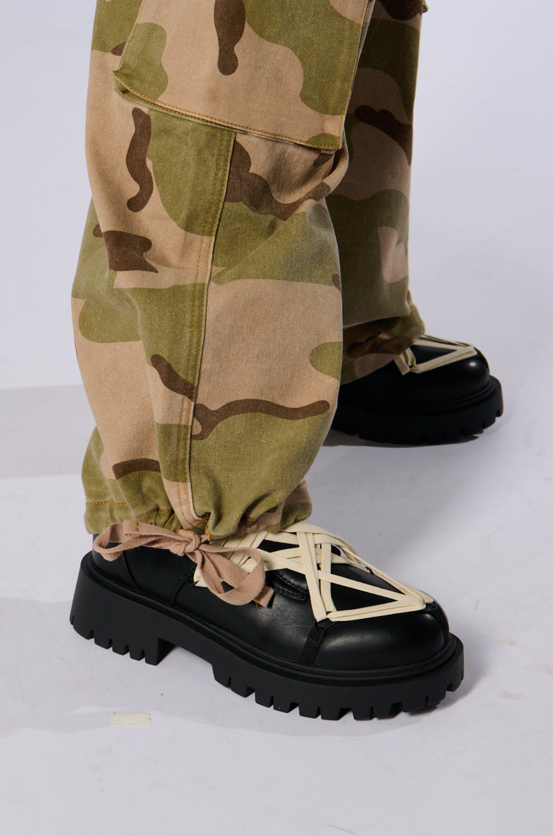 Close-up of a person wearing the ON TOP OF IT CAMO JOGGER pants with cargo flap pockets and black platform shoes. The jogger pants are gathered at the ankles with drawstrings, and the shoes feature crisscrossing beige laces. The background is plain white.