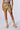 Model showcasing the ON TOP OF IT CAMO SHORT, featuring a high-rise fit with multiple buckles and cargo pockets, paired with a cropped top and beige chunky boots worn with socks. Visible tattoos adorn the midriff and thigh, set against a plain, neutral background.