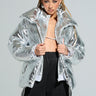 A person with braided hair wears a SPACE CASE METALLIC PUFFER jacket and black pants featuring white stripes. They are looking confidently at the camera, standing against a plain background.