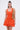 A woman wears the LET'S GO TO THE BEACH RUFFLE MINI DRESS IN ORANGE—sleeveless with a textured skirt—styled with gold earrings and a tan woven shoulder bag, standing against a plain light background.