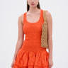 A woman wears the LET'S GO TO THE BEACH RUFFLE MINI DRESS IN ORANGE—sleeveless with a textured skirt—styled with gold earrings and a tan woven shoulder bag, standing against a plain light background.