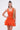A woman stands against a white background wearing the LET'S GO TO THE BEACH RUFFLE MINI DRESS IN ORANGE, a sleeveless, textured mini dress with a ruffled skirt, and holds a woven tan bag over her shoulder—showcasing the perfect summer look.