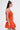 A woman with long, straight brown hair models the LET'S GO TO THE BEACH RUFFLE MINI DRESS IN ORANGE, featuring a flared skirt. Styled in profile with gold statement earrings and a woven bag against a white background—ideal for summer.