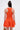 A woman with long dark hair is shown from the back wearing the LET'S GO TO THE BEACH RUFFLE MINI DRESS IN ORANGE. The sleeveless, fitted mini dress features a ruffled skirt. She holds a woven bag and has a medical device on her upper arm.