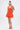 A woman poses against a plain background in the LET'S GO TO THE BEACH RUFFLE MINI DRESS IN ORANGE, featuring a textured, ruffled skirt—ideal for summer—styled with tan platform sandals and a woven tan shoulder bag.