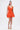 A woman stands against a white background, wearing the LET'S GO TO THE BEACH RUFFLE MINI DRESS IN ORANGE with ruffled details and a flared skirt. She carries a woven bag, wears lace-up platform sandals, and has her hair in a high ponytail.
