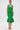 A woman faces forward in the LET'S GO TO THE BEACH RUFFLE MIDI DRESS IN GREEN, a sleeveless fitted dress with a textured bodice and ruffled hem. She accessorizes with hoop earrings, neutral makeup, and lace-up wedge sandals against a plain white background.