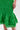 A close-up shows a person wearing the LET'S GO TO THE BEACH RUFFLE MIDI DRESS IN GREEN, featuring textured ruched fabric and an eyelet ruffle hem. Their hand is at their side, with part of their bare leg and foot visible—ideal for summer style.