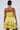 A person with their hair in a sleek bun is shown from the back, wearing the Ready For Summer Ruffle Mini Dress in yellow eyelet fabric and boasting a ruffled hem. They are holding a metallic silver clutch in their right hand.