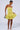 A woman stands against a plain background, showcasing the READY FOR SUMMER RUFFLE MINI DRESS—a strapless, yellow, ruffled mini dress. She holds a small silver handbag in her right hand and wears white sandals. Her hair is styled back, and she looks down with a soft smile.