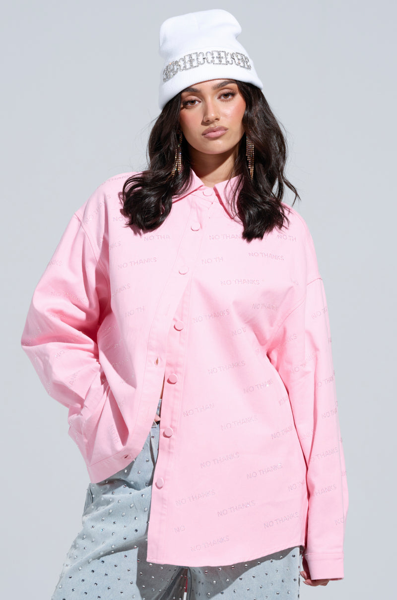 A person wears a pink NO THANKS BUTTON DOWN TOP with subtle text, paired with light grey pants featuring small patterns. Their long hair cascades beneath a white beanie embellished with rhinestones, and large hoop earrings complete the look as they pose against a plain background.