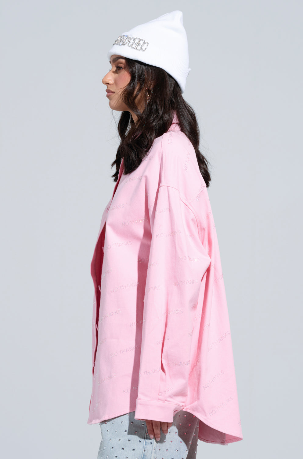 A person wearing the NO THANKS BUTTON DOWN TOP in long sleeves and pink, along with a white beanie, is seen in profile against a plain background. Their hair is dark, and they are wearing a light blue, patterned skirt.