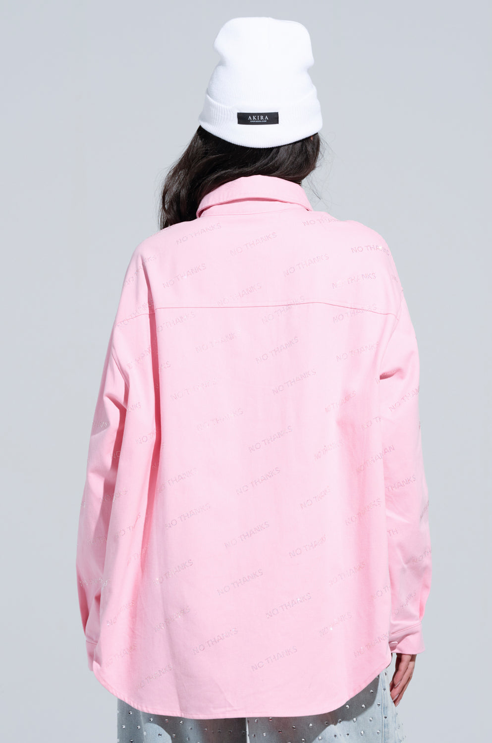 A person wearing a white beanie and an oversized NO THANKS BUTTON DOWN TOP, printed with "No Thanks," stands with their back to the camera against a light gray background, giving off a casual and laid-back vibe.