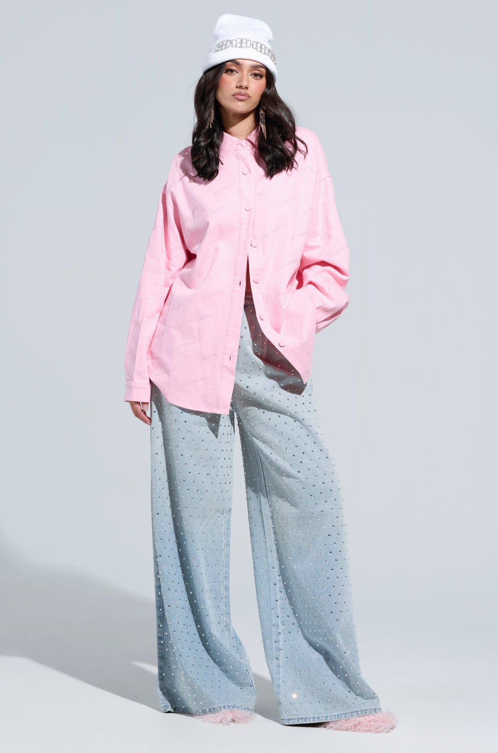 A person wearing a NO THANKS BUTTON DOWN TOP in pink and light blue wide-leg pants embellished with rhinestones stands against a light gray background. They have long dark hair and a white knit bucket hat.
