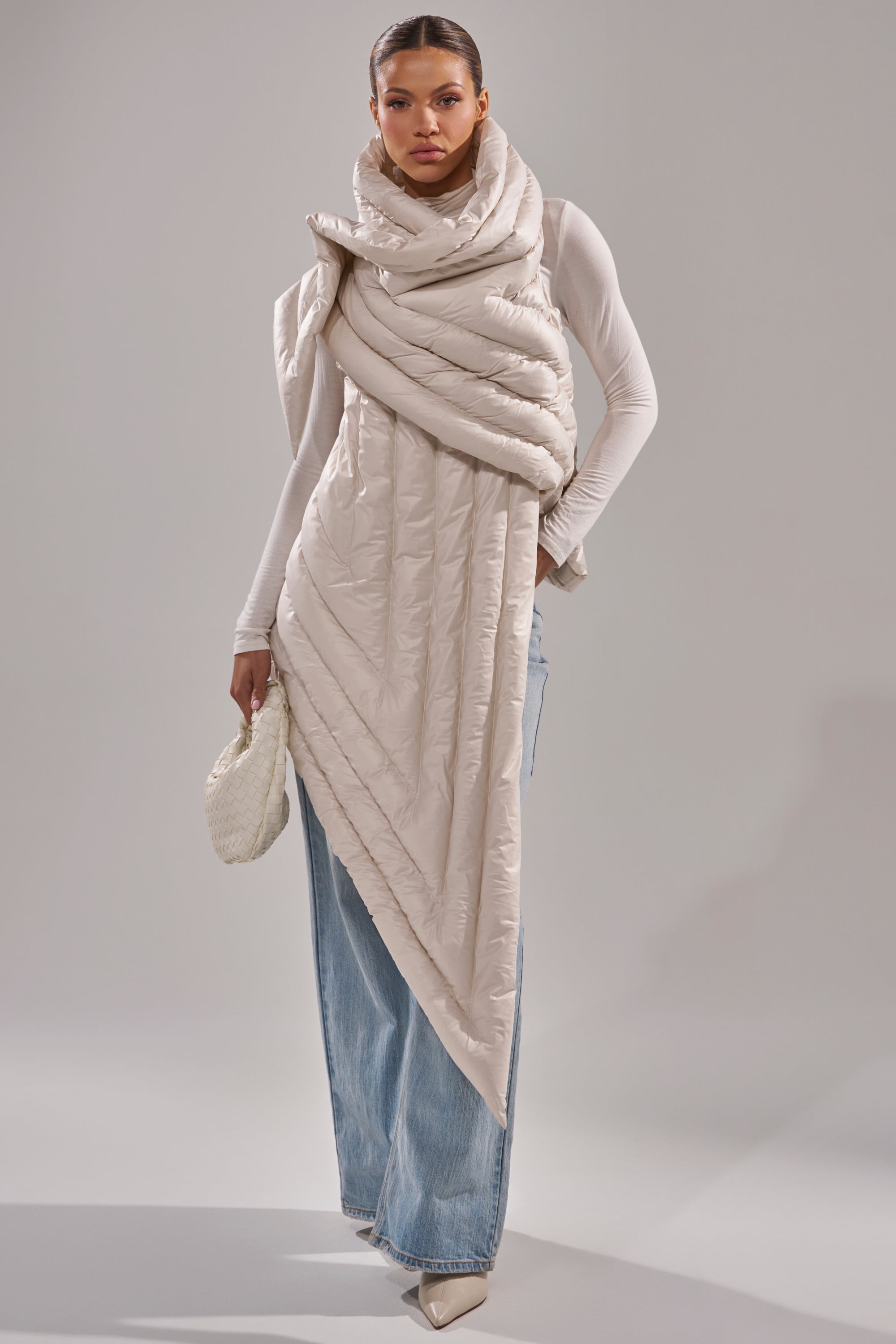 A woman styles the FOR THE DRAMA PUFFER VEST IN IVORY over a fitted white top and wide-leg light blue jeans, accessorized with a textured white clutch and pointed-toe shoes, posed against a plain background. 0