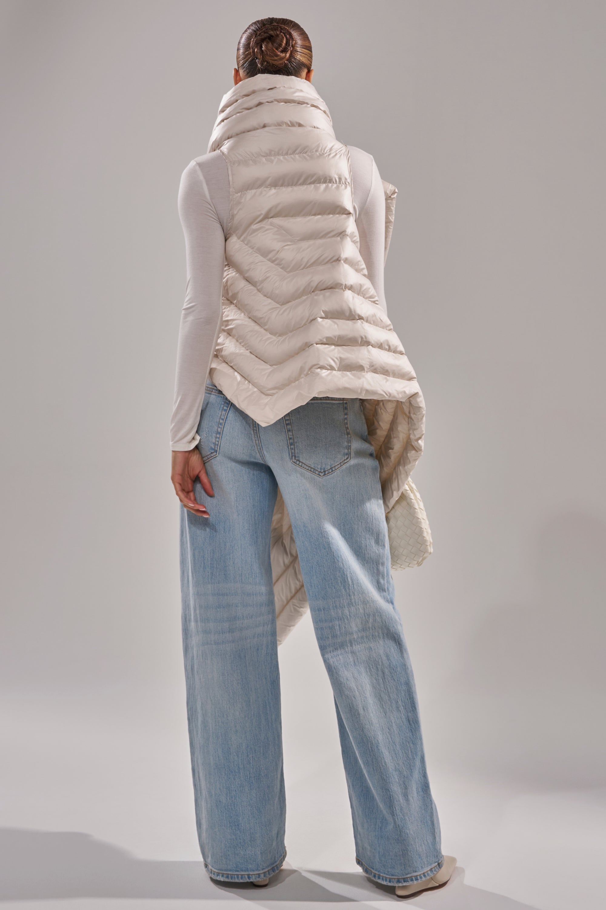 A person with dark hair in a bun faces away, wearing the FOR THE DRAMA PUFFER VEST IN IVORY over a white long-sleeve top and loose light blue jeans, set against a plain, light-colored background. 1