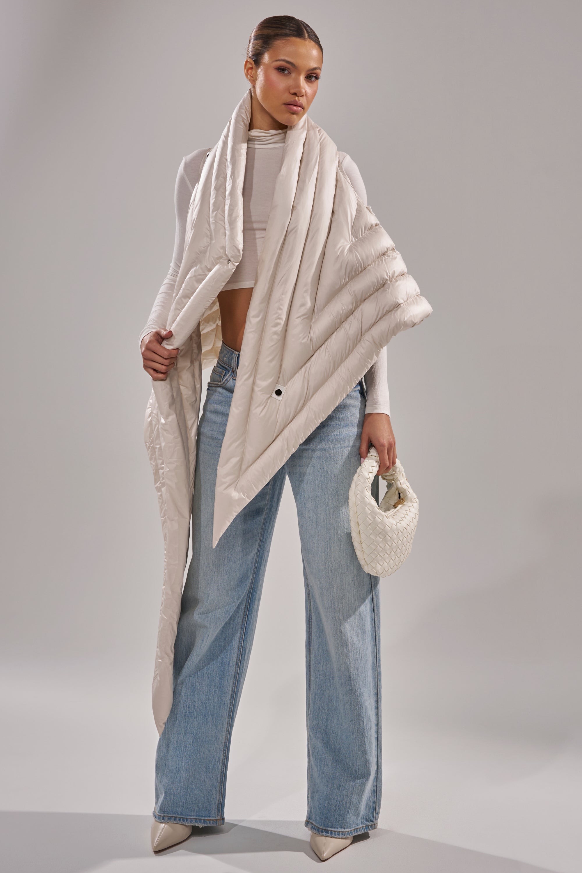 A woman stands in a studio wearing the FOR THE DRAMA Puffer Vest in Ivory, paired with a white cropped turtleneck, wide-leg light blue jeans, pointed-toe heels, and holding a small woven white handbag. 2