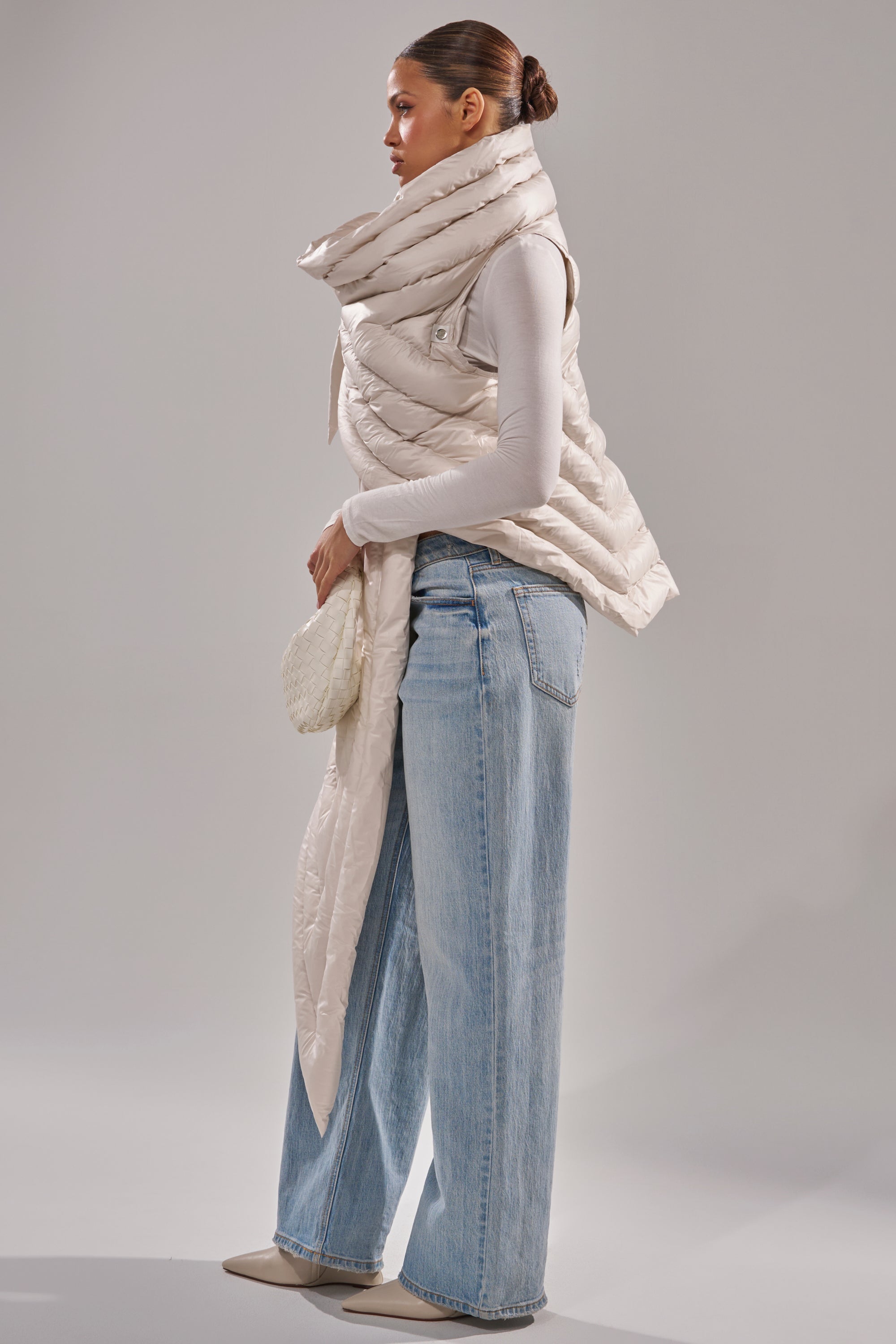 A woman stands sideways wearing the FOR THE DRAMA PUFFER VEST IN IVORY, featuring a long scarf-like collar and asymmetrical hem, paired with a fitted white turtleneck, light blue wide-leg jeans, beige pointed shoes, and a cream clutch. 3