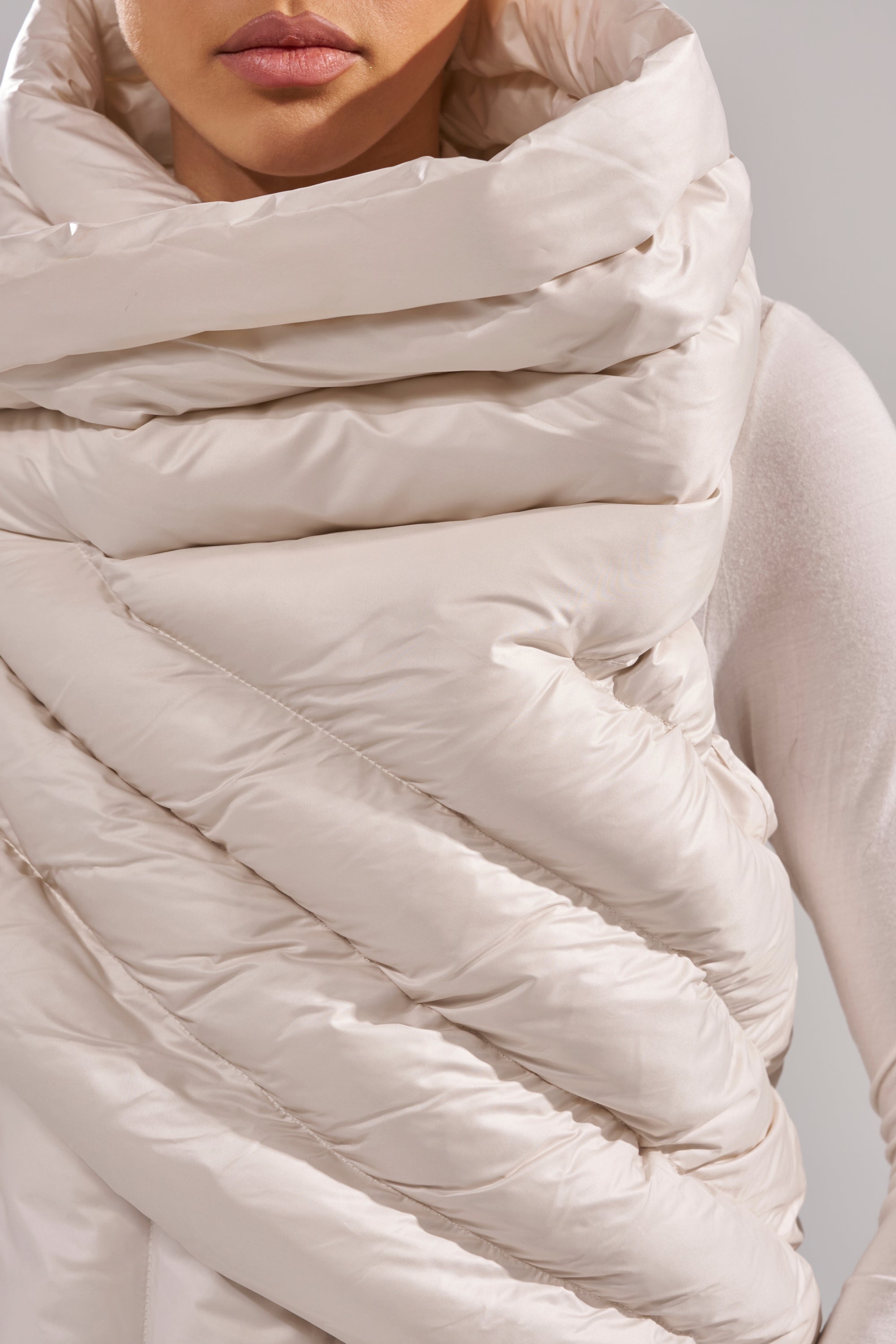 A person wearing the FOR THE DRAMA PUFFER VEST IN IVORY, a thick, shiny, quilted vest with a wrap-around design in cream, styled to cover their neck and shoulders and partially conceal their face. 5