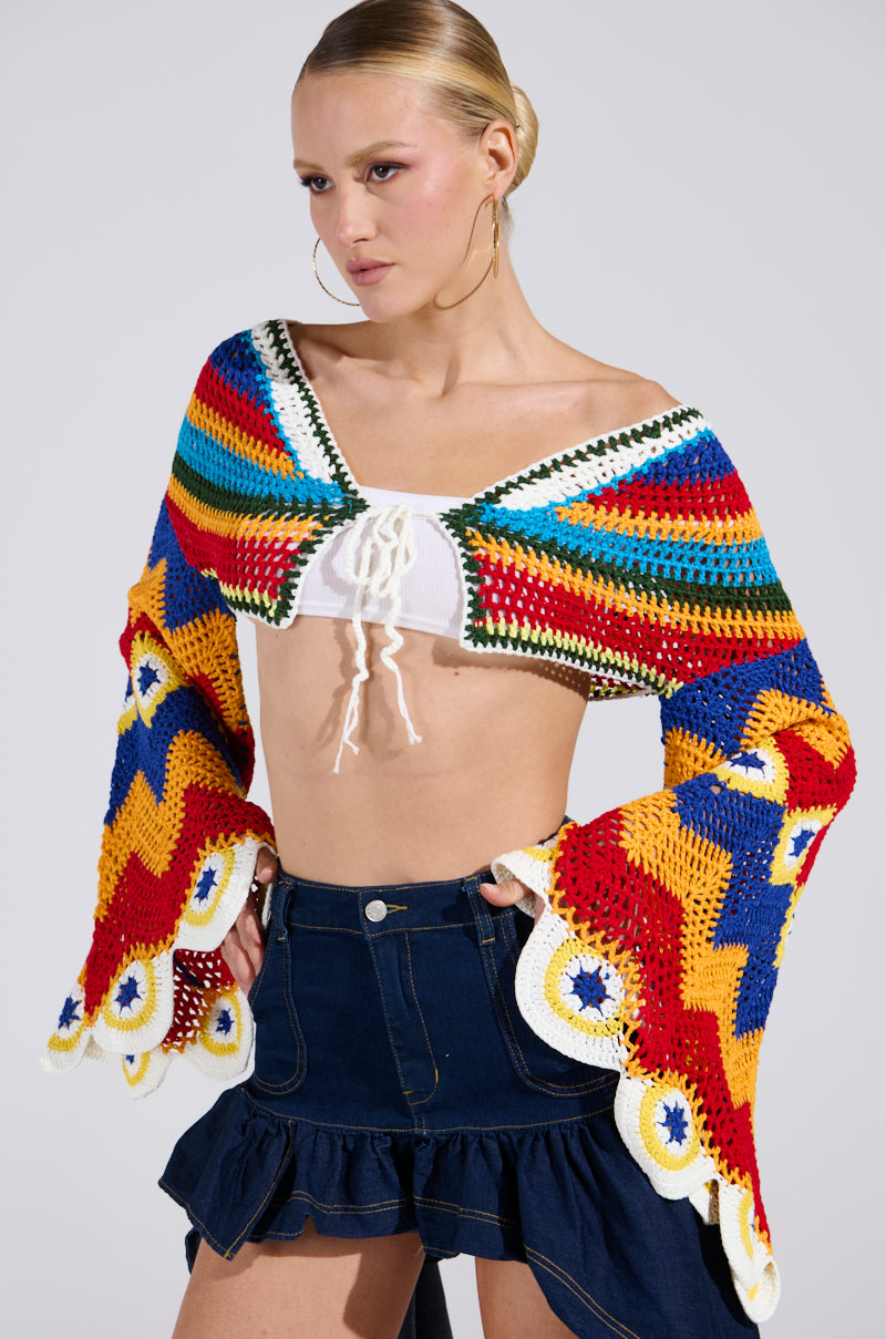 A person pairs a WARM DAYS CROCHET TOP featuring red, blue, yellow, and green patterns with bell sleeves over a white top. They accessorize with hoop earrings and style their blond hair back, complementing a dark denim skirt.