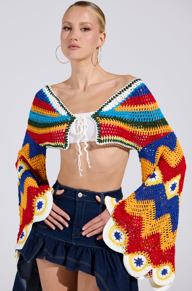 A person confidently poses in the WARM DAYS CROCHET TOP, a colorful zigzag-patterned shawl in vibrant red, yellow, blue, green, and orange over a white plunging neckline top, paired with a dark denim skirt and large hoop earrings.