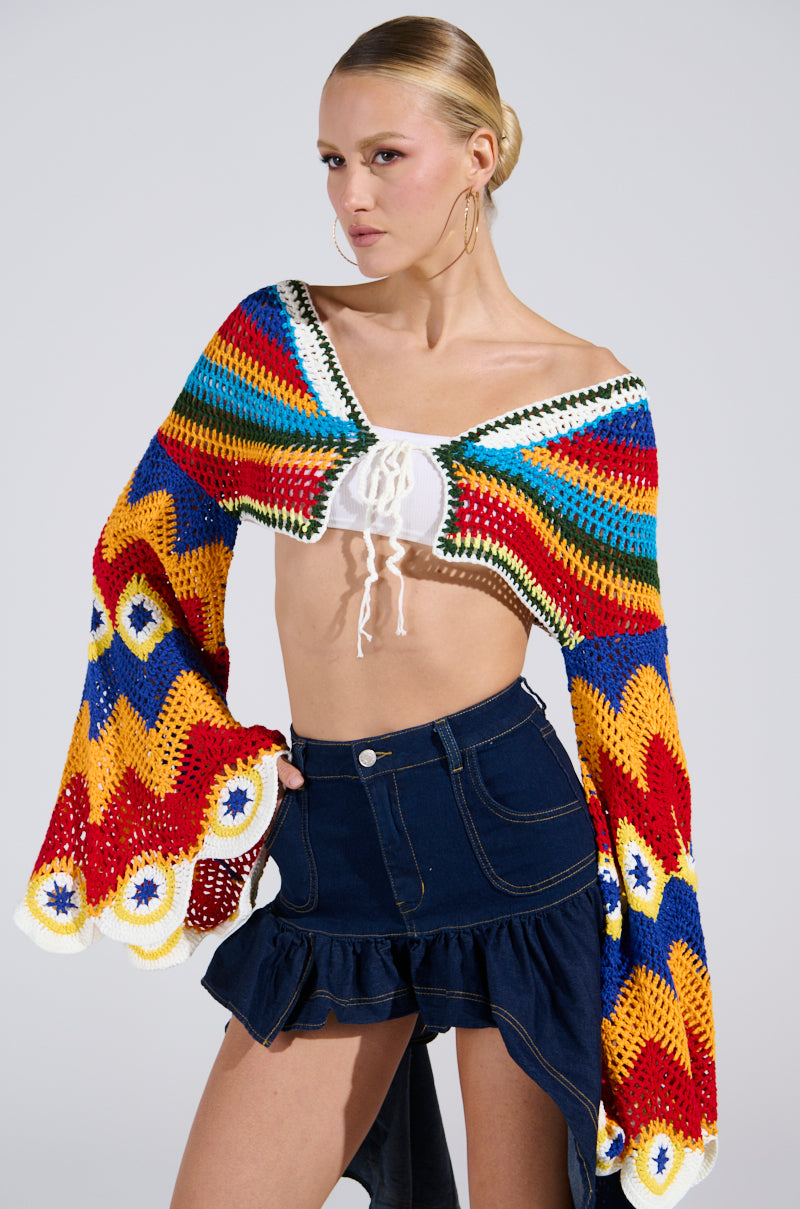 They don a colorful zigzag crocheted wrap, a plunging neckline white WARM DAYS CROCHET TOP, and a dark blue denim skirt with ruffled edges against a plain light gray background.