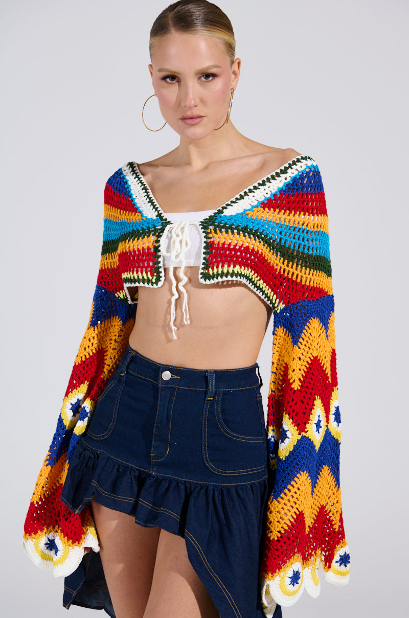 A person with slicked-back hair wears the WARM DAYS CROCHET TOP, showcasing red, blue, yellow, and green patterns, over a white crop top with bell sleeves. They pair it with a dark blue denim skirt featuring ruffled layers and finish the look with large hoop earrings.