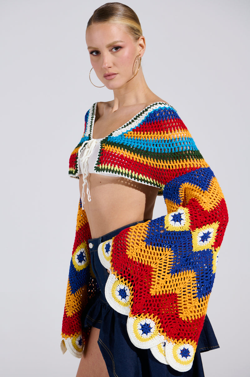 A model confidently poses in the WARM DAYS CROCHET TOP, featuring a plunging neckline and large bell sleeves with intricate geometric patterns in red, yellow, green, and blue. She sports bold hoop earrings and gazes directly at the camera against a plain background.