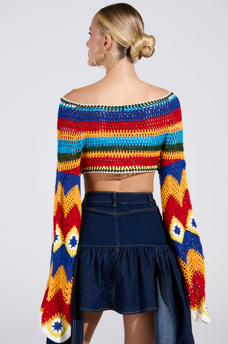 A blonde-haired person with a bun wears the vibrant WARM DAYS CROCHET TOP featuring off-the-shoulder, striped zig-zag patterns, and bell sleeves. It’s paired with a denim skirt with a ruffled hem against a light gray background.