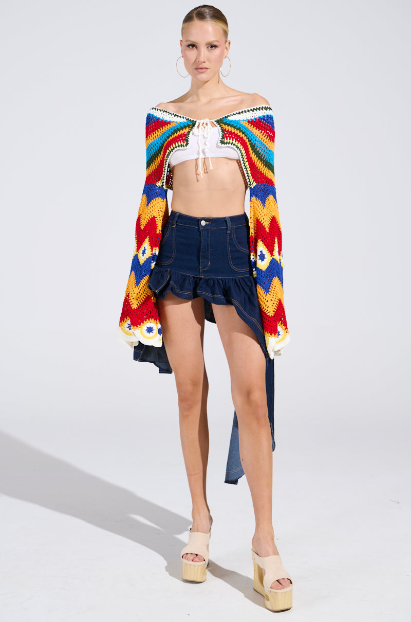 A woman stands against a light background wearing the colorful WARM DAYS CROCHET TOP, which features an off-the-shoulder design and geometric patterns with flared bell sleeves. She pairs it with a white cropped lace-up blouse, dark high-waisted ruffled shorts, platform heels, hoop earrings, and a sleek hairstyle.