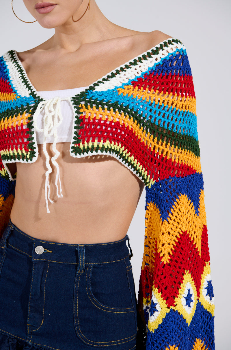 Clad in the WARM DAYS CROCHET TOP, featuring a plunging neckline, vibrant patterns, and bell sleeves, a person pairs it with high-waisted dark denim jeans. Only part of their face is visible.