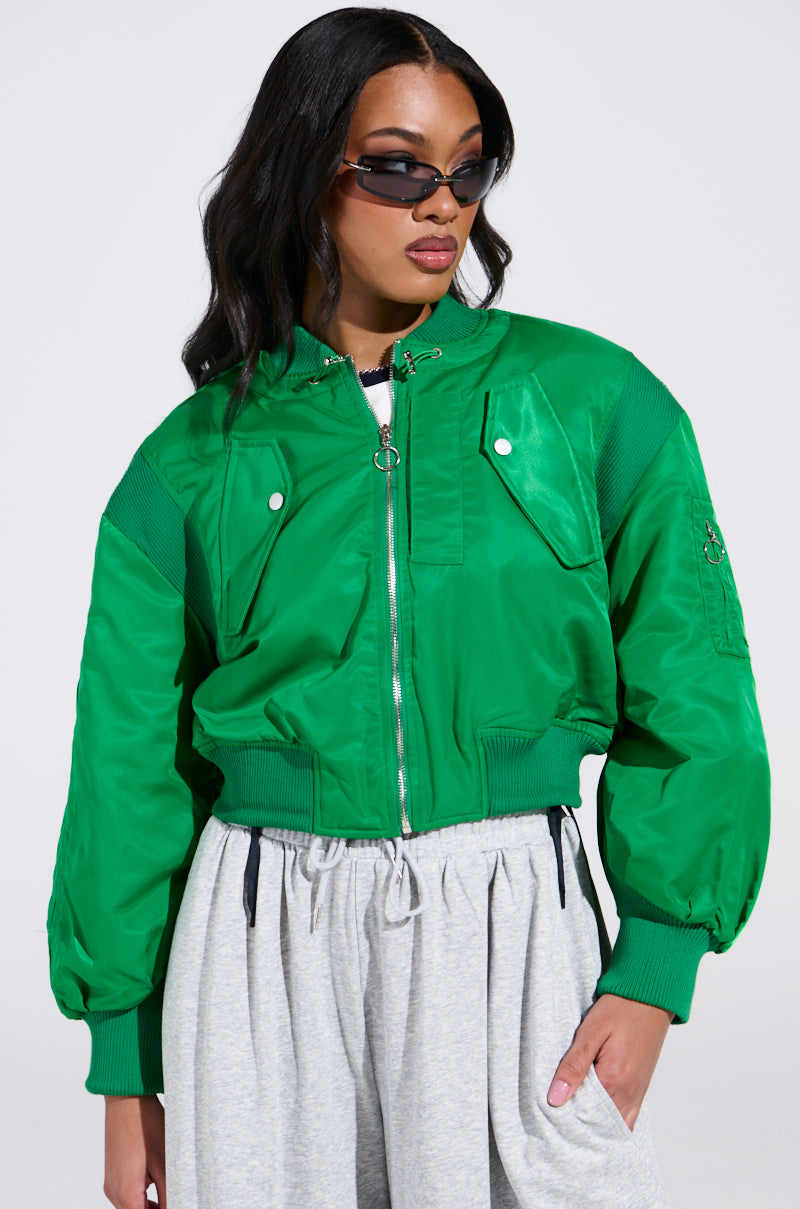 A person confidently poses in the LUCKY ME ASYMMETRICAL BOMBER IN GREEN, gray sweatpants, and dark sunglasses. They have long, dark hair and are set against a plain white background.