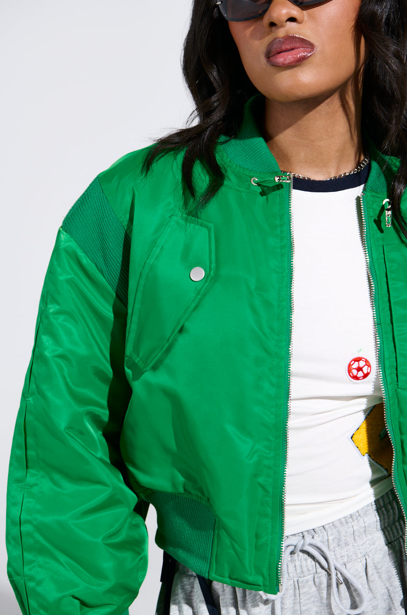 The woman sports the LUCKY ME ASYMMETRICAL BOMBER IN GREEN over a white shirt featuring a red and yellow graphic, paired with gray sweatpants. Her long dark hair and sunglasses complement her look, emphasizing the unique bomber's windbreaker fabrication.