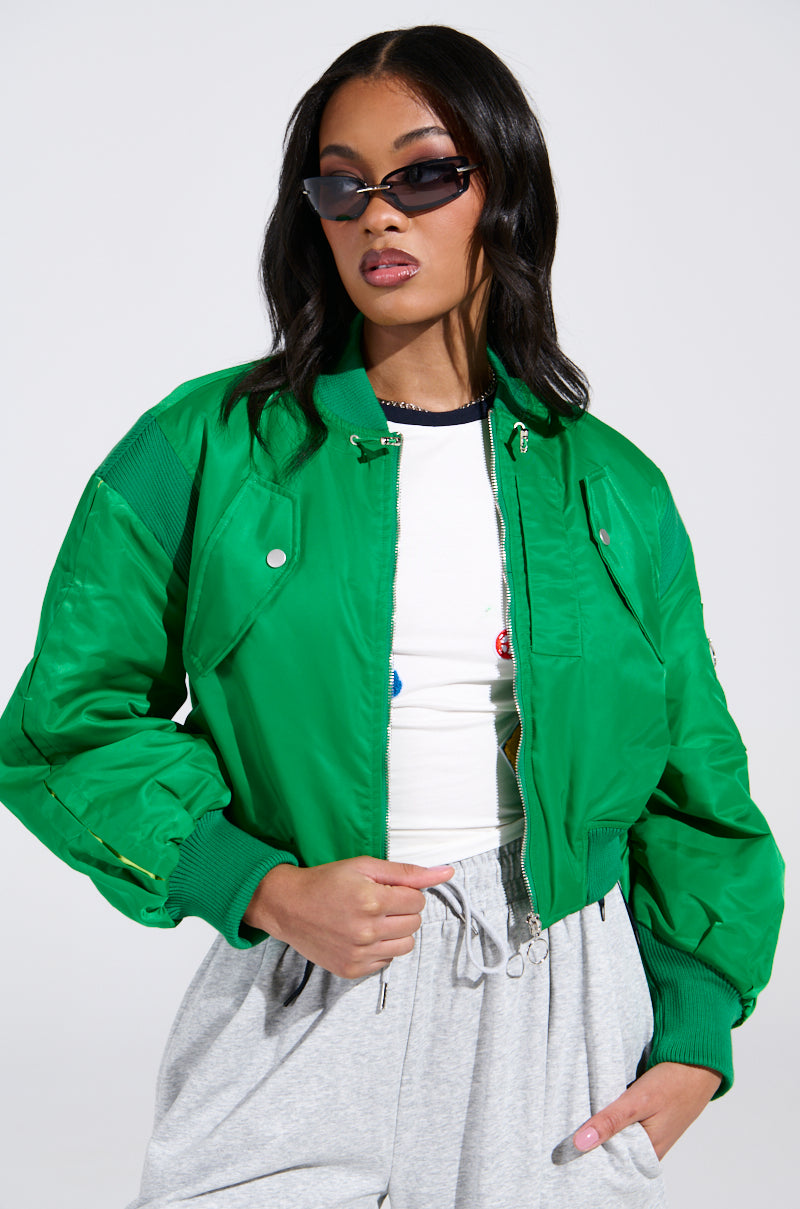Wearing long hair, a person sports the LUCKY ME ASYMMETRICAL BOMBER IN GREEN over a white shirt with colorful buttons, paired with gray sweatpants and chic sunglasses, confidently posing against a plain white background.