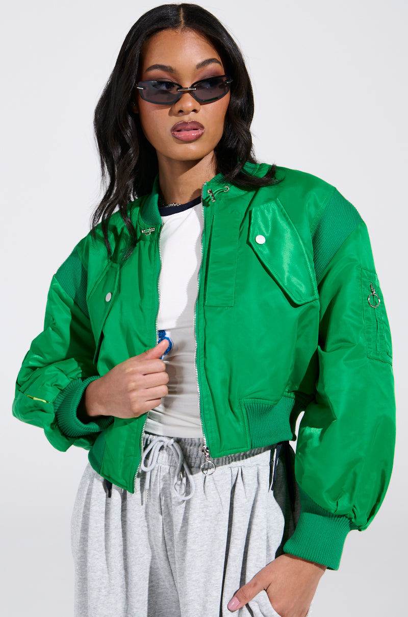 A person wearing the LUCKY ME ASYMMETRICAL BOMBER IN GREEN over a white shirt and gray sweatpants. With long dark hair and black sunglasses, they grip the jacket with one hand while keeping the other in their pocket, set against a plain white background.