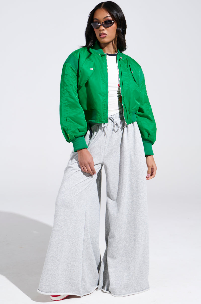 A woman confidently poses in the LUCKY ME ASYMMETRICAL BOMBER IN GREEN, paired with a white top and wide-legged gray pants. She sports sunglasses and pink shoes, standing against a plain white background.