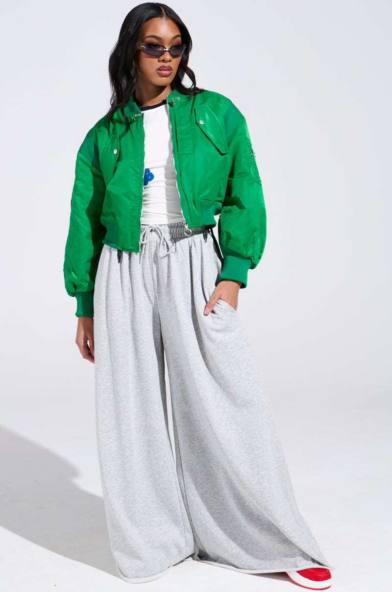 Dressed in the LUCKY ME ASYMMETRICAL BOMBER IN GREEN, a person with long dark hair and sunglasses exudes confidence. They pair the jacket with a white shirt, oversized gray wide-legged pants, and white sneakers featuring touches of red.