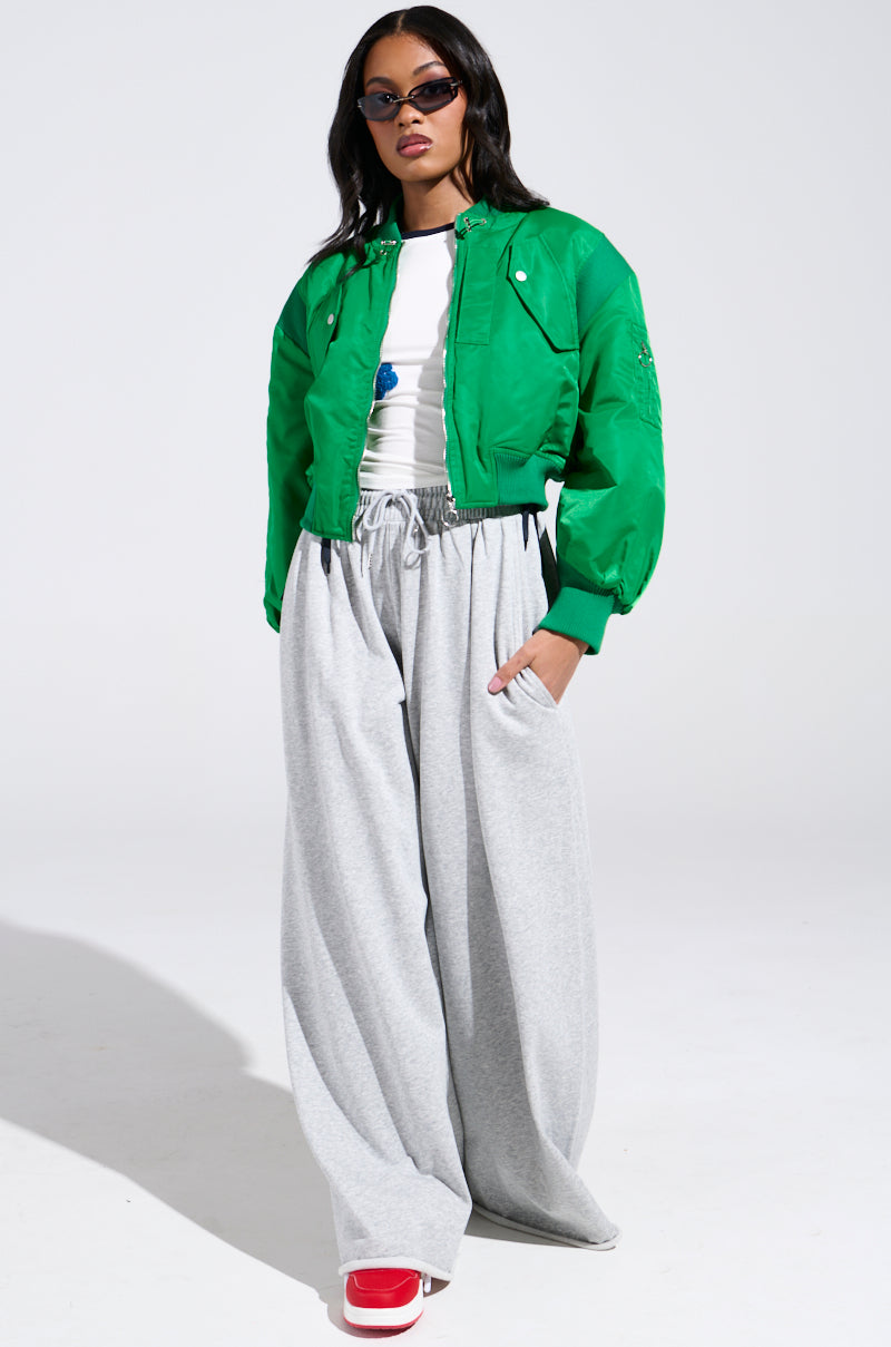 A person stands confidently against a plain background, wearing the LUCKY ME ASYMMETRICAL BOMBER IN GREEN over a white shirt, paired with gray wide-leg sweatpants and vibrant red sneakers. With long hair and sunglasses, they exude effortless style.