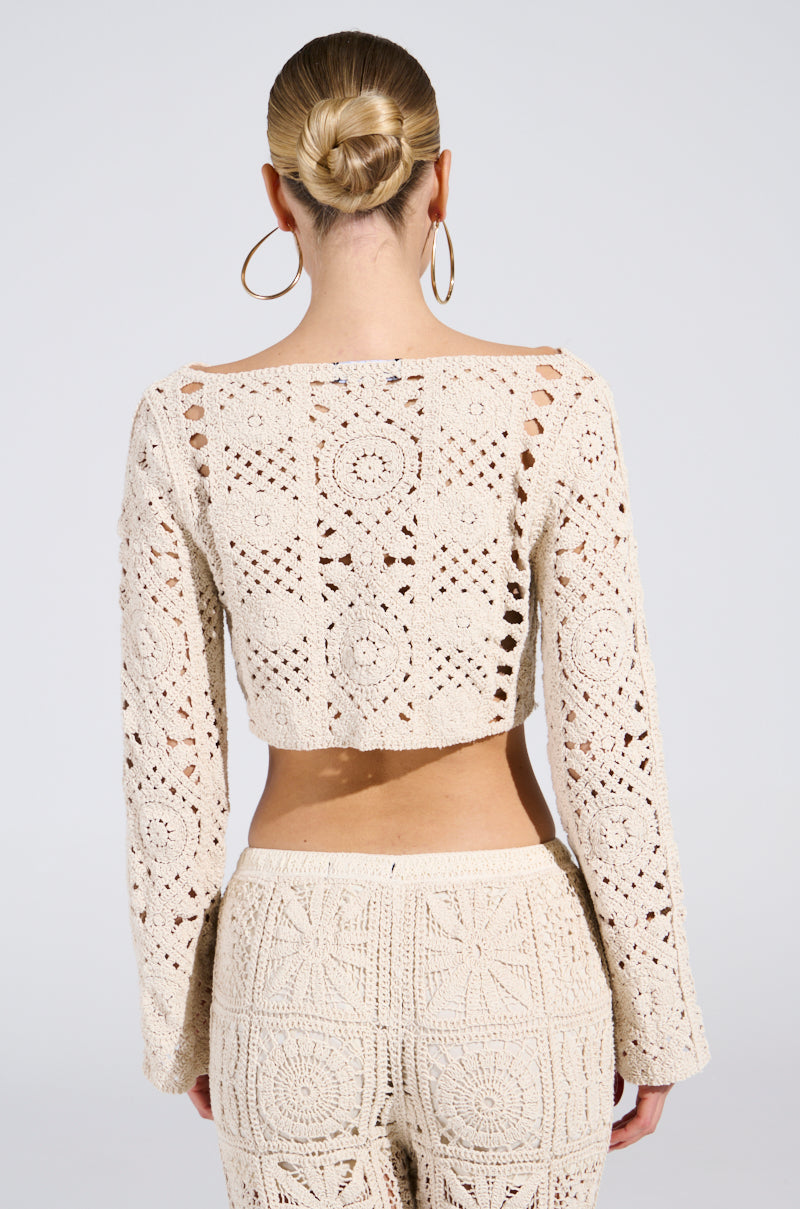 A person with a bun is wearing a CROCHET KNIT CROP TOP and matching pants, cream-colored with intricate circular and geometric patterns. The sun-kissed vibe outfit is shown from the back to highlight its design.
