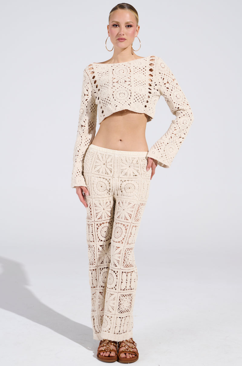 Wearing the CROCHET KNIT CROP TOP and matching pants with intricate square patterns, styled with large hoop earrings and brown sandals, they exude a sun-kissed vibe against a plain white background.