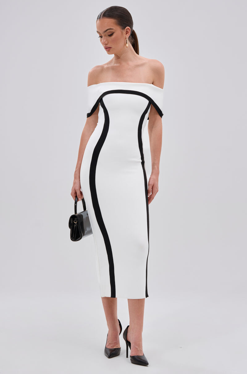 A woman poses against a plain light background in the SLEEK AND CHIC BANDAGE MIDI DRESS, featuring a white off-the-shoulder design with black curved lines, paired with black pointed heels and a small black handbag.