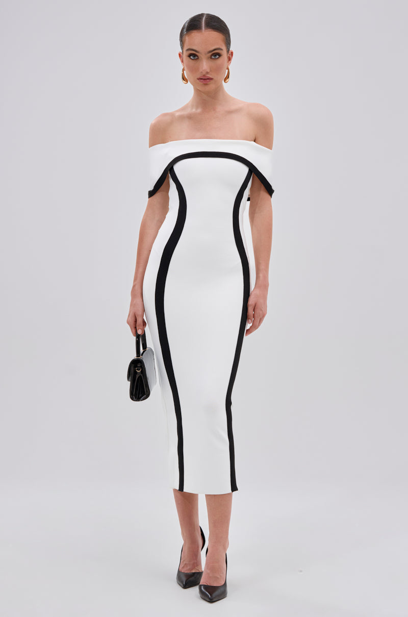 A woman in the SLEEK AND CHIC BANDAGE MIDI DRESS with an off-the-shoulder neckline, black heels, gold hoop earrings, and a black handbag stands against a plain light background.