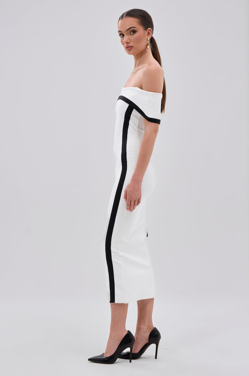 A woman stands sideways in the SLEEK AND CHIC BANDAGE MIDI DRESS, featuring a bodycon fit, off-the-shoulder neckline, white fabric with black vertical stripes, and black high heels against a plain light gray background.