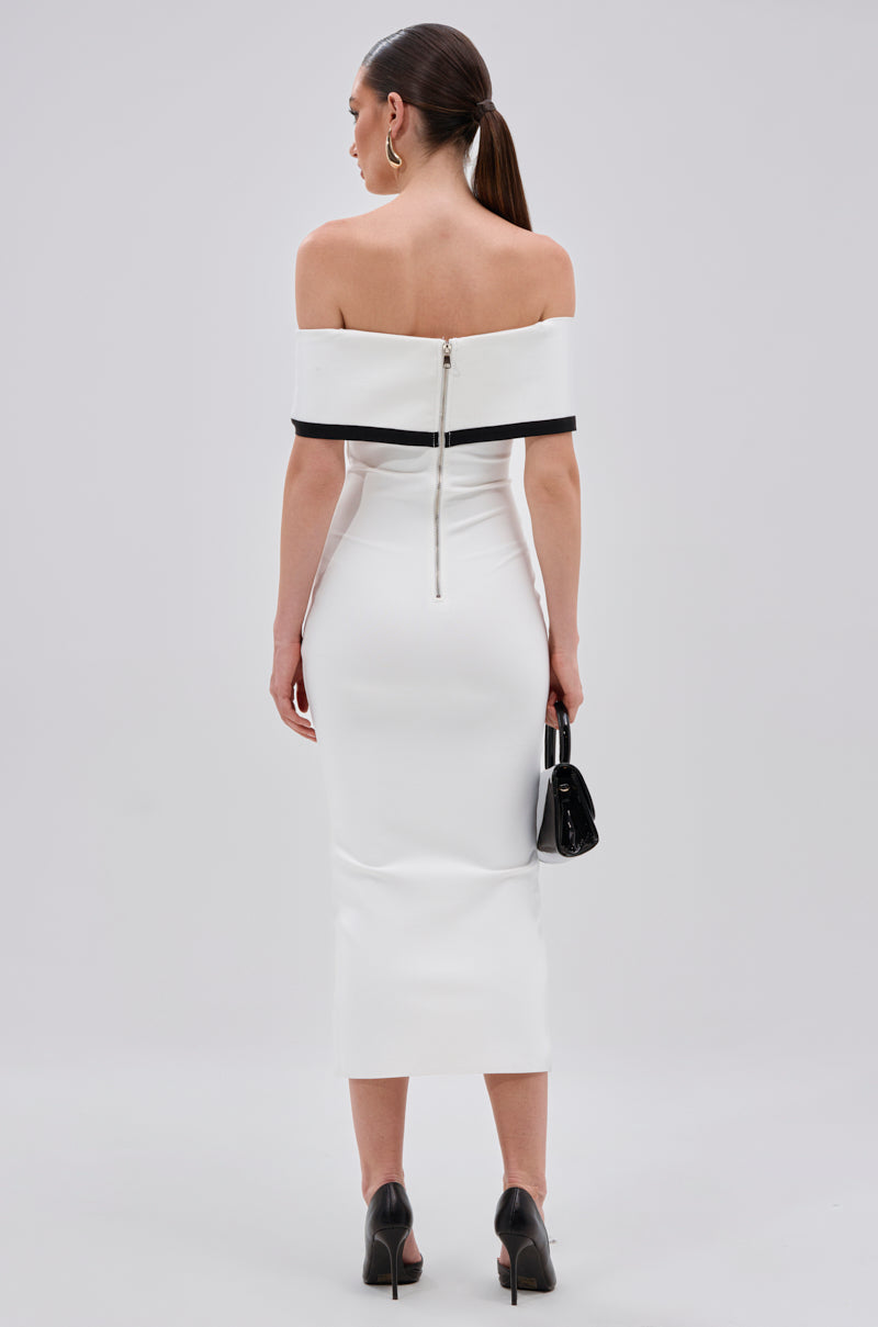 A woman faces away, wearing the SLEEK AND CHIC BANDAGE MIDI DRESS—an off-the-shoulder white bodycon midi with black trim and a back zipper. She completes the look with black heels, a small black handbag, and a sleek ponytail.
