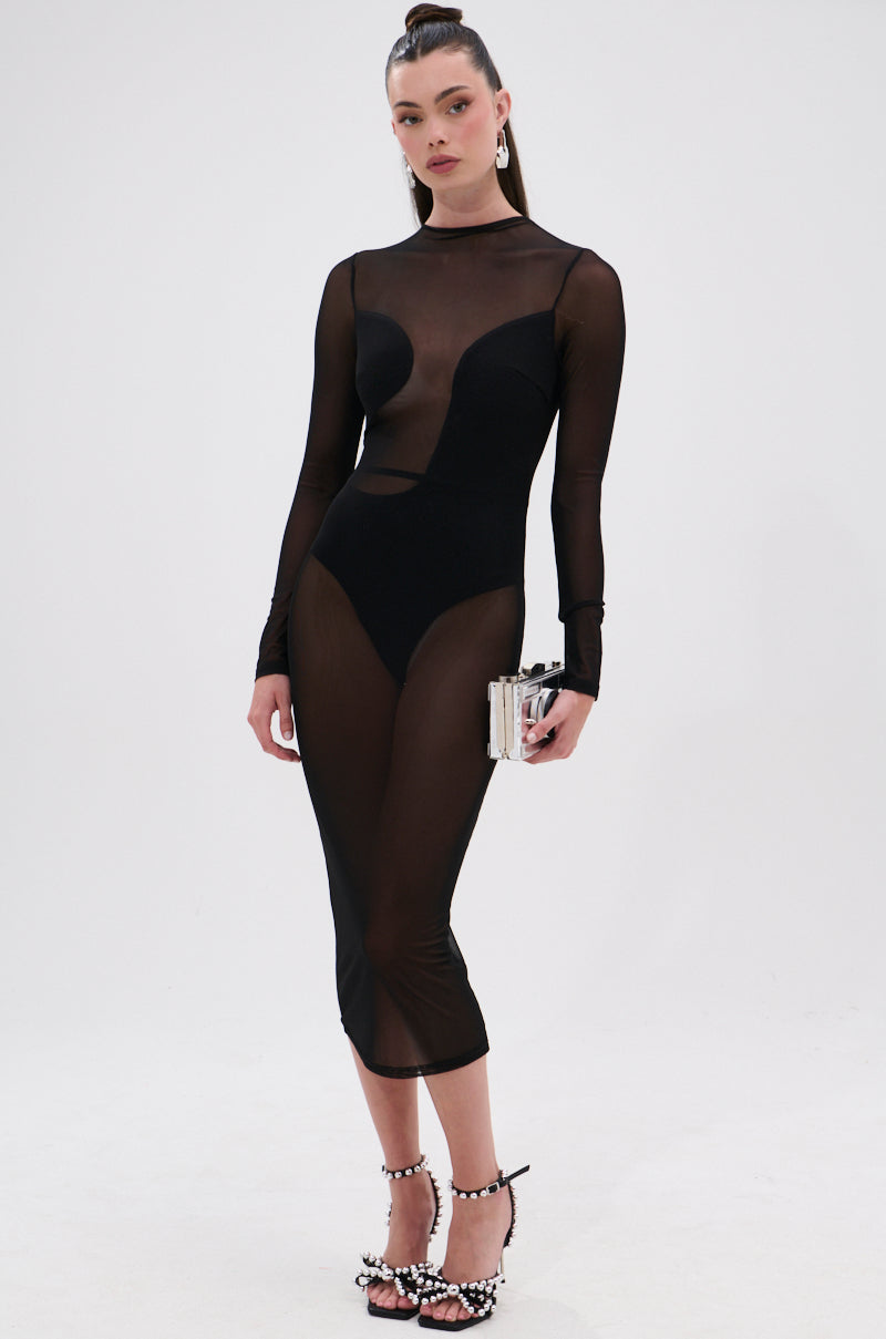 A woman poses against a white backdrop wearing the MINDING MY BUSINESS MESH MIDI DRESS over a black bodysuit, accessorized with pearl-embellished heels, statement earrings, a high ponytail, and carrying a small clutch.
