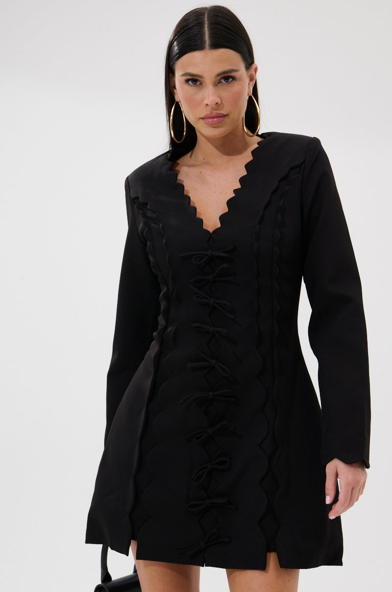 A woman with straight dark hair wears the CUTE AS CAN BE MINI DRESS WITH BOWS, which features a scalloped V-neckline and decorative bows down the front. She pairs it with large hoop earrings and carries a small black handbag.