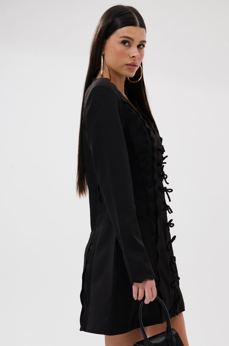 A woman with long straight dark hair stands against a plain light background wearing the CUTE AS CAN BE MINI DRESS WITH BOWS, gold hoop earrings, and holding a small black handbag.