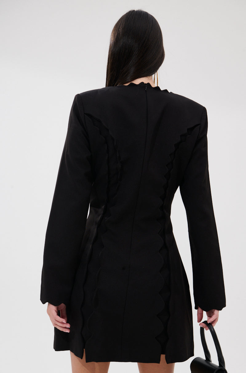 A woman with long straight hair, seen from behind, wears the CUTE AS CAN BE MINI DRESS WITH BOWS—a fitted black mini dress with wavy trim and long sleeves—while holding a small black handbag against a light background.